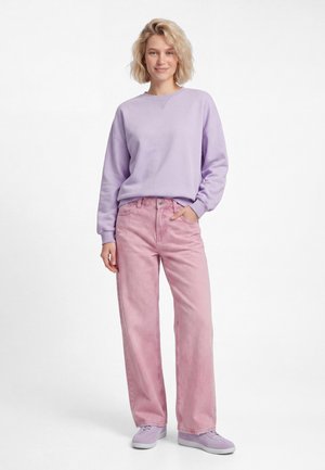 Lachende vrouw met kort blond haar draagt een lila trui, roze wijd uitlopende jeans en lila sneakers, staat met één hand in haar zak.