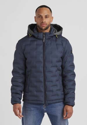 Marineblaue gepolsterte Jacke mit Kapuze, ausgestattet mit einer gesteppten Textur, einem Reißverschluss vorne und grünen Kordeln. Getragen über blauen Jeans.