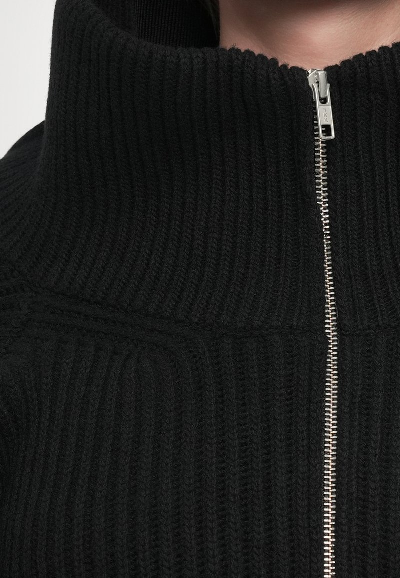 Chaqueta de punto negra con un alto cuello y detalle de cremallera plateada. Presenta una textura acanalada y un diseño estructurado. Vista en primer plano del escote.