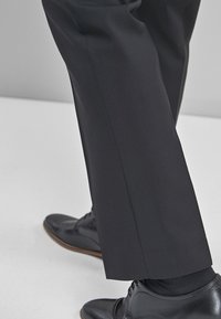 Schwarze maßgeschneiderte Hose mit geradem Schnitt und Bügelfalte. Kombiniert mit schwarzen Lederschuhen, die eine glatte, polierte Oberfläche zeigen.