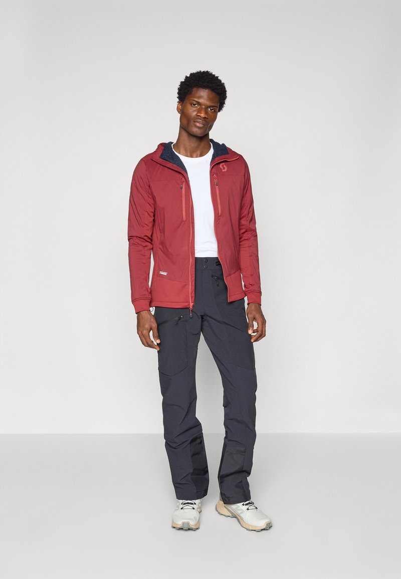 Rode waterdichte jas met een ritssluiting aan de voorkant, capuchon en twee met rits afgesloten zakken, gecombineerd met donkere broek en lichte sneakers.