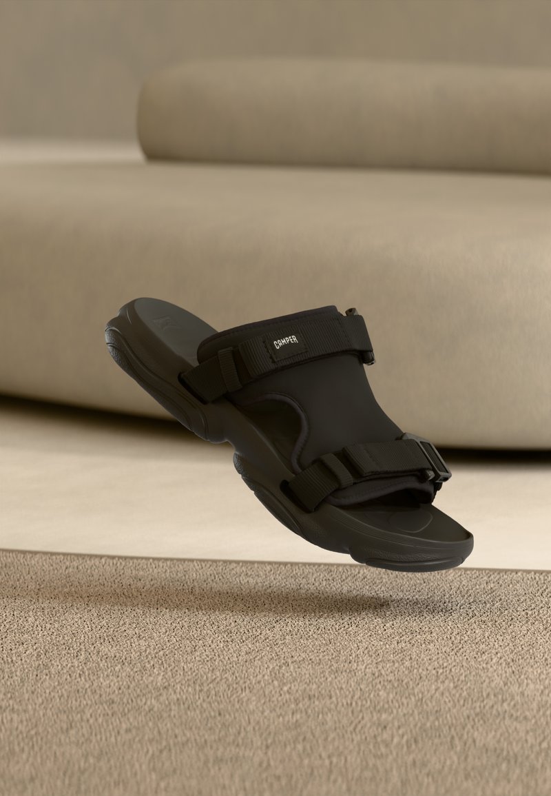 Sandale à bride noire réglable avec semelle épaisse flottant au-dessus d'un tapis beige, canapé beige doux en arrière-plan.