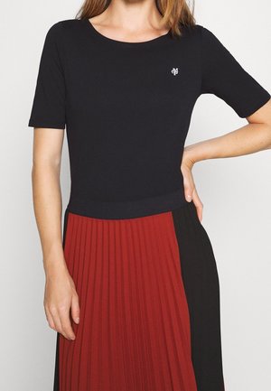 Woman wearing a black short-sleeve top with a small white logo and a pleated skirt with red and black vertical panels.