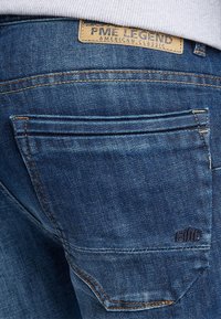 Vaqueros de denim con un lavado azul oscuro, que cuentan con un bolsillo trasero, detalles de costura y un parche de logo de cuero. Textura suave con un ligero brillo.