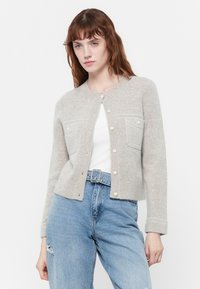 Ung kvinne iført en lys grå knappet strikkegenser over en hvit topp og høymidjede blå jeans med et belte med sølvspenne.
