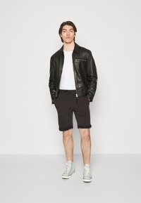 Veste en cuir noire avec fermeture éclair à l'avant, t-shirt blanc, shorts noirs et baskets montantes bleu clair. Le modèle se tient dans un cadre neutre.