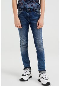 Slim fit modré džíny s vybledlými místy a pěti kapsami, nošené s bílými teniskami s černými akcenty a texturovanou podrážkou.