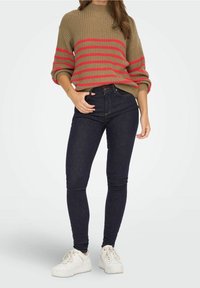 Suéter marrón de punto con rayas horizontales rojas, combinado con jeans ajustados oscuros y zapatillas blancas. El suéter presenta un cuello alto y mangas con puños.