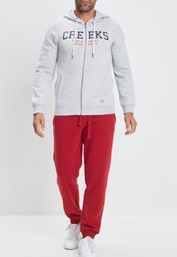 Sudadera gris con cremallera y logo, con cordones blancos y bolsillos, combinada con pantalones de chándal rojos y zapatillas blancas sobre un fondo simple.