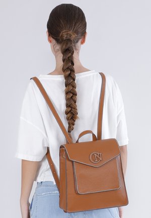 Persona con capelli lunghi intrecciati che indossa una camicia bianca e jeans azzurri chiari, con uno zaino di pelle marrone con dettagli a fibbia.