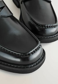 Schwarze Lederschuhe mit glänzender Oberfläche, ausgestattet mit einer genähten Spitze und einer dicken schwarzen Gummisohle. Nahaufnahme, die Textur und Handwerkskunst hervorhebt.