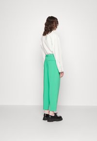 Persoon die zijwaarts staat in felgroene 3/4-broek, wit langetermijn shirt en zwarte chunky veterschoenen tegen een effen achtergrond.