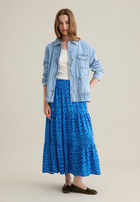 Giacca di jeans blu chiaro con chiusura a bottoni e taschino sul petto, abbinata a una gonna maxi a strati blu con fantasia, e scarpe basse scure.