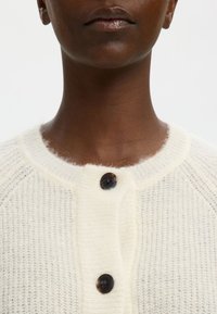 Off-white adīta jaka ar apaļu kakla izgriezumu, ar trim tumšiem pogām un mīkstu, teksturētu apdari. Pamanāmi raglanu piedurknes.
