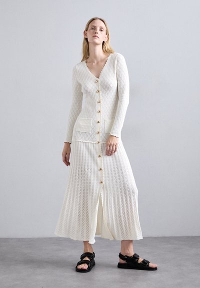 Witte gebreide lange cardigan met een V-hals, gouden knopen en twee voorzakken. Gestructureerd patroon met een bijpassende geplooide rok. Zwarte sandalen.
