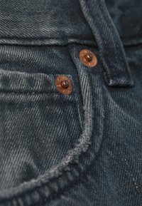 Detailní záběr tmavě modré džínoviny ukazující roh kapsy se dvěma měděnými nýty a detailním prošitím.