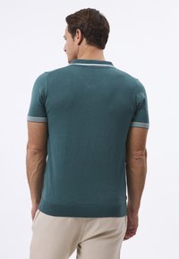 Teal värvi lühikeste varrukatega polo särk ribidega krae ja triibuliste aktsentidega varrukatel, valmistatud siledast, kergest kangast. Näha on heledbežžid püksid.