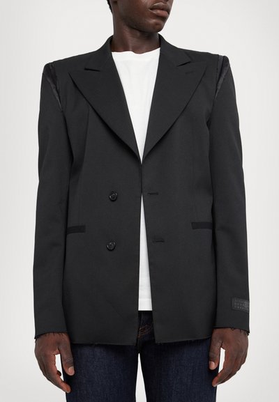 Blazer noir à double boutonnage avec des revers pointus, en tissu lisse, deux boutons et des ourlets effilochés. Il est associé à un t-shirt blanc en dessous et un jean foncé.