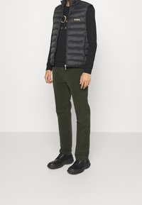 Gilet imbottito nero con colletto alto, logo dorato, abbinato a una camicia a maniche lunghe nera, pantaloni verde oliva e scarpe nere con suola texturizzata.