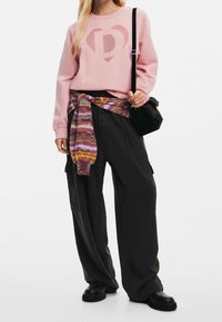 Rosa sweatshirt med ett prickigt hjärtmotiv, svarta vida byxor och en flerfärgad randig scarf knuten i midjan. Svart axelväska och skor.