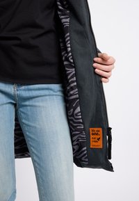 Graue Jacke mit Reißverschluss und einem gemusterten schwarz-grauen Innenfutter, ausgestattet mit einem orangen Etikett mit der Aufschrift "Wir sind eins. Peace Clothing." und einer strukturierten Oberfläche.