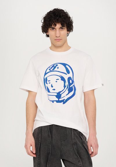 Λευκό βαμβακερό t-shirt με γραφικό αστροναύτη σε μπλε χρώμα. Κοντά μανίκια, στρογγυλή λαιμόκοψη και άνετη εφαρμογή. Φορεμένο με γκρι υφασμάτινο παντελόνι.