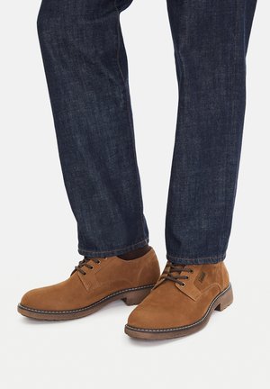 Brązowe zamszowe buty o gładkiej fakturze, z okrągłym noskiem i sznurowadłami. Do tego ciemnoniebieskie dżinsy z mankietem na dole.