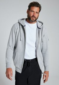 Felpa grigia con zip e coulisse regolabili, indossata sopra una t-shirt bianca. Pantaloni neri con vita a coulisse.