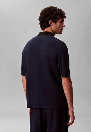 Άνδρας με σγουρά μαλλιά που φοράει μια κοντομάνικη μπλε σκούρα μπλούζα με μαύρο γιακά, στέκεται γυρισμένος με την πλάτη σε ένα απλό γκρι φόντο.