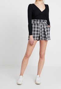Schwarzes langärmliges Wickeltop, kombiniert mit hochtaillierten Karshorts mit einem Bindegürtel in Schwarz, Weiß und Grau, und weißen Sneakers.