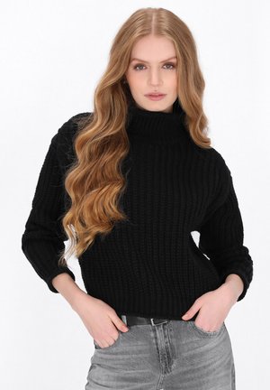 Ung kvinde med langt bølget lyst hår iført en sort striktrøje og grå jeans, stående med hænderne i lommerne foran en hvid baggrund.