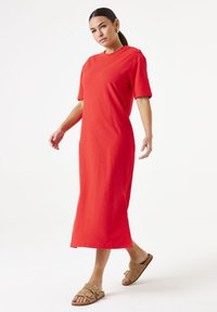 Vestido midi de algodón rojo con mangas cortas, escote redondo y detalle de abertura lateral. Combinado con sandalias marrones tipo slide con hebillas.