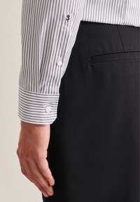 La mano sinistra di un uomo accanto a pantaloni neri e una manica di camicia a righe verticali grigio e bianca con polsino abbottonato e dettaglio ricamato di una rosa.