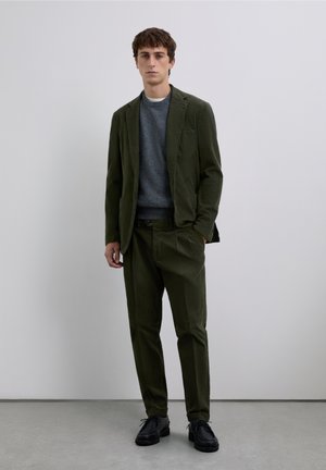 Chaqueta de traje de pana verde oliva y pantalones a juego con un suéter gris de cuello redondo. Zapatos negros con cordones completan el conjunto.