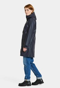 Marinblå vattentät regnkappa med huva, med rak passform, två sidofickor och justerbara ärmslut, i kombination med blå jeans och svarta stövlar.