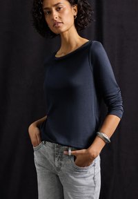 Navyblauwe, langemouwen top in een gladde stof, gecombineerd met lichtgrijze jeans. Kenmerkt zich door een bateauhals en een zilveren armbandaccent.