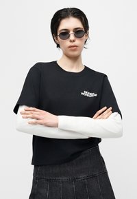 Άτομο με μαύρα γυαλιά ηλίου, μαύρο κοντομάνικο πουκάμισο πάνω από λευκό μακρυμάνικο πουκάμισο και σκοτεινή πλισέ φούστα, με σταυρωμένα τα χέρια.