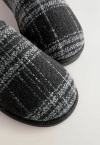 Mustat ja harmaat ruudulliset tohvelit, jotka on valmistettu pehmeästä huovasta, pyöreällä kärjellä ja teksturoidulla mustalla kumipohjalla pidon takaamiseksi.