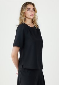 Kobieta o blond włosach, ubrana w czarny top z krótkim rękawem i pasującą spódnicę, stojąca bokiem na jednolitym jasnym tle.