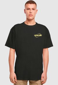 T-shirt en coton noir avec un col rond, présentant un logo "upscale" jaune sur la poitrine gauche. Manches courtes et légèrement ample.