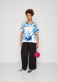 Satinbluse med blå blomsterprint, sorte plisserte bukser, metalliske blå flate sko og en rund rosa teksturert håndveske.