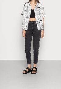 Kurzärmliges, weißes Hemd mit schwarzen Cartoon-Drucken, kombiniert mit hochgeschnittenen schwarzen Jeans und schwarzen flachen Sandalen mit gekreuzten Riemen.