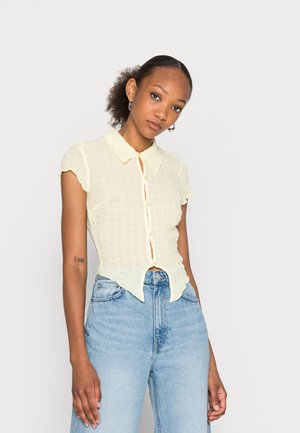 Camisa de manga curta com colarinho de cor amarelo-claro, com um padrão texturizado, fecho de botão e uma bainha ligeiramente curva, combinada com jeans azuis de cintura alta.