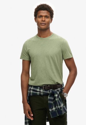 Camiseta de manga corta color verde oliva con cuello redondo, combinada con pantalones verde oscuro y una camisa de cuadros azul atada a la cintura, complementada con un reloj.