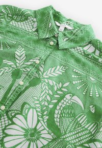 Chemise verte à boutons avec des motifs tropicaux blancs détaillés comprenant des feuilles, des fleurs, un oiseau et un col à motifs.
