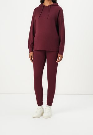 Personne portant un sweat à capuche bordeaux assorti et un jogging slim avec des baskets blanches, debout devant un fond blanc uni.