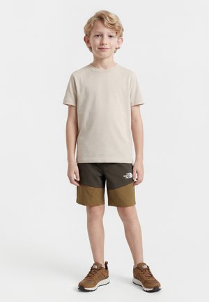 Jongen staat, draagt een beige T-shirt, bruin tweekleurige korte broek en bruine sneakers, tegen een effen witte achtergrond.
