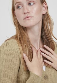 Cardigan côtelé vert olive, avec un col en V et une fermeture à boutons, en tissu doux et texturé. Des mains touchent délicatement l'encolure.
