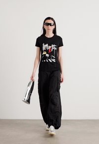 Must musta mustriga T-särk, millel on värviline trükk, ja mustad cargo-püksid. Modell hoiab läikivat hõbedast kotti ning kannab tumedaid prille.