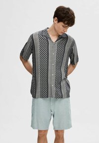 Camicia a maniche corte, con motivo in bianco e nero, con scollo a V e bottoni, abbinata a pantaloni corti azzurri. Il tessuto appare leggero e texture.